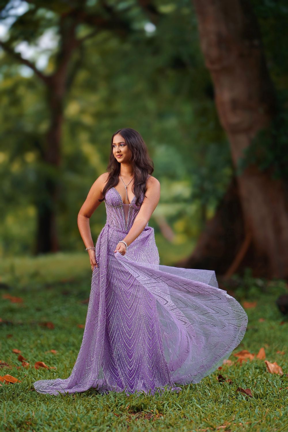 A person in a flowing lavender gown poses gracefully on grass in an outdoor garden setting.