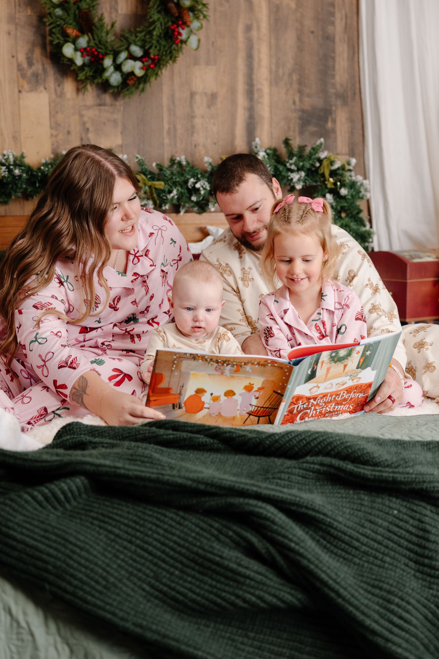 Family wearing matching Christmas pajamas reads a storybook together on bed with green blanket and holiday decorations.