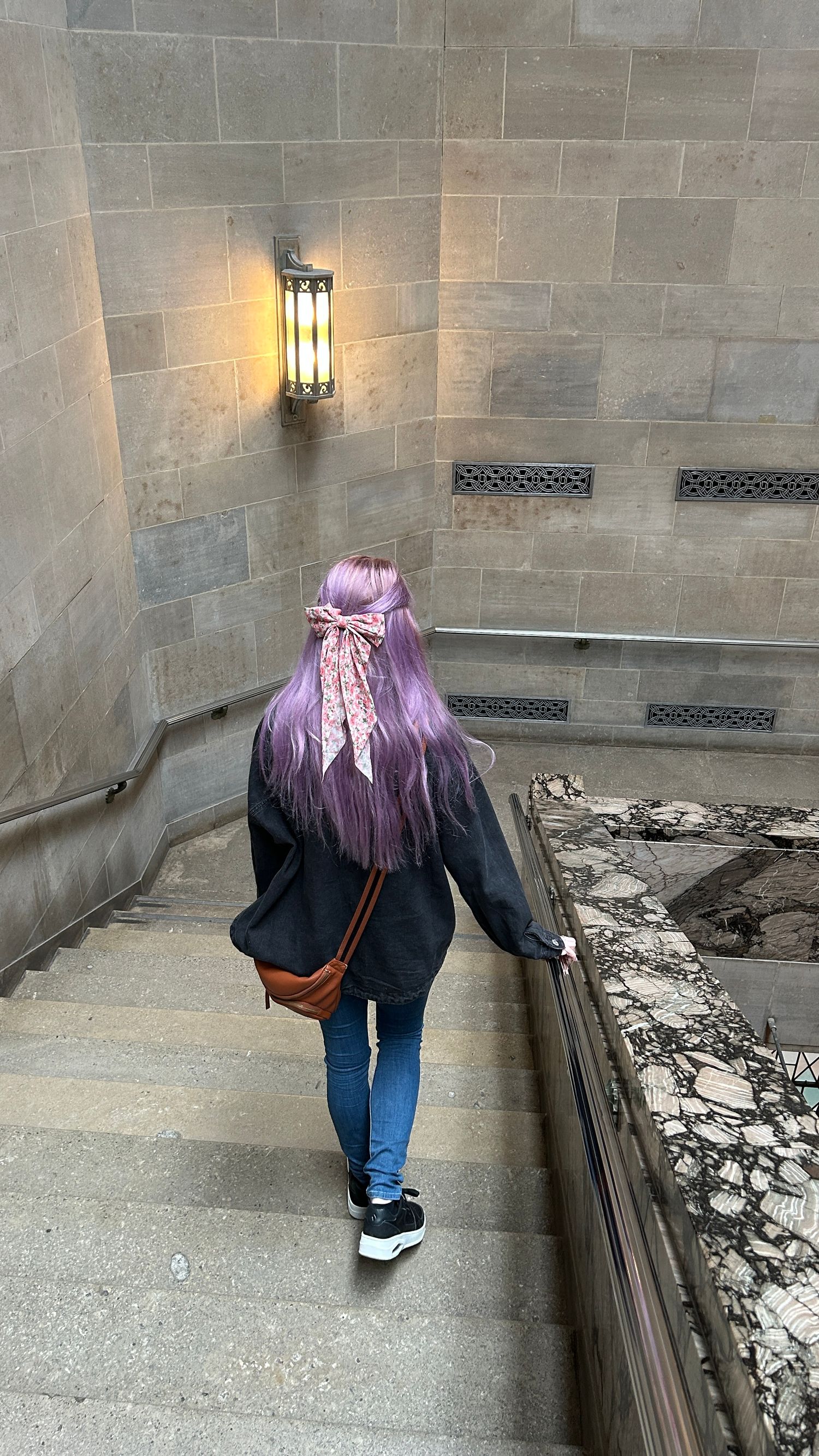 Person with purple hair descending stone stairs in a medieval castle corridor with wall-mounted lantern.
