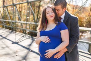 A couple in coordinating blue and black outfits embrace during an outdoor autumn photoshoot on a wooden bridge.