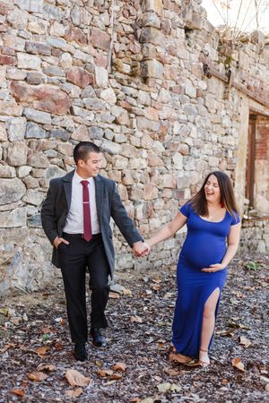 A couple in formal attire poses playfully against a rustic stone wall during a photo session.
