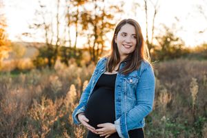 Expectant mother in denim jacket poses in golden field at sunset with gentle smile.