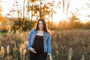 A person in a denim jacket and black dress stands in a golden field at sunset with warm autumn lighting.