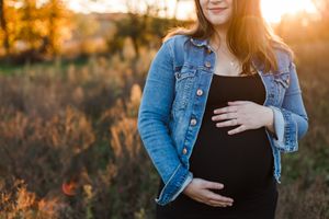 Close up of pregnant belly with hands resting on it, wearing denim jacket at sunset.