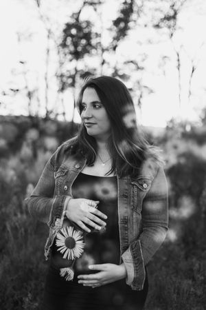 Black and white maternity photo showing woman in denim jacket standing in field.