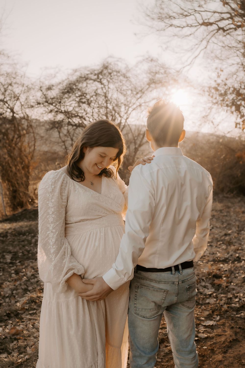 A couple stands together in a sunlit forest setting during golden hour, with the woman wearing a flowing white dress.