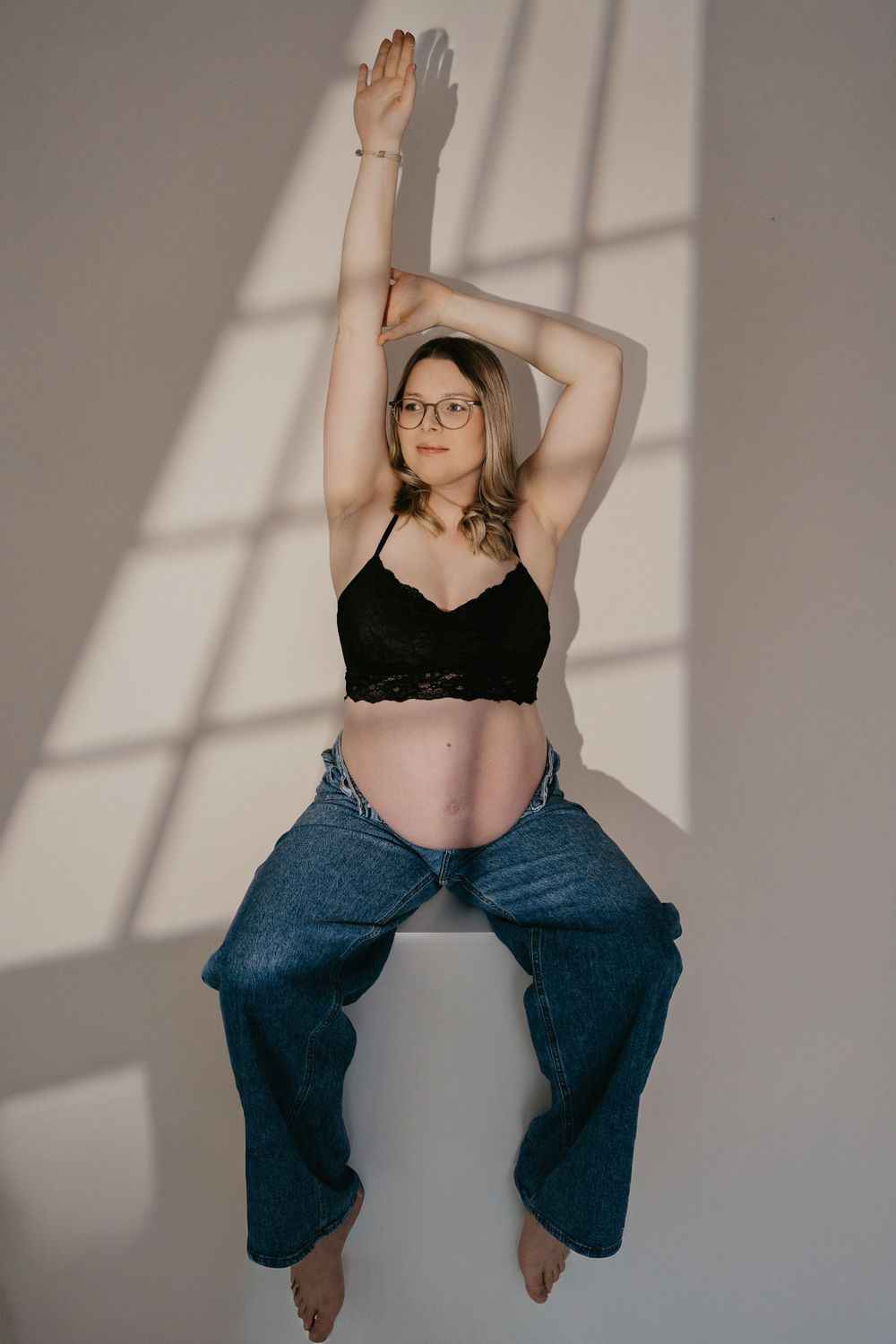 Artistic boudoir photo series showing person in black bralette and jeans against white wall with window shadows.
