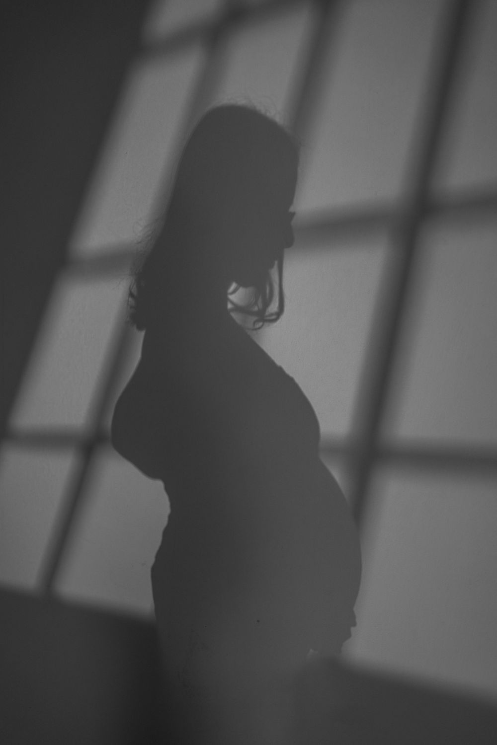 Silhouette of a pregnant woman standing in dramatic window light casting shadows on the wall.