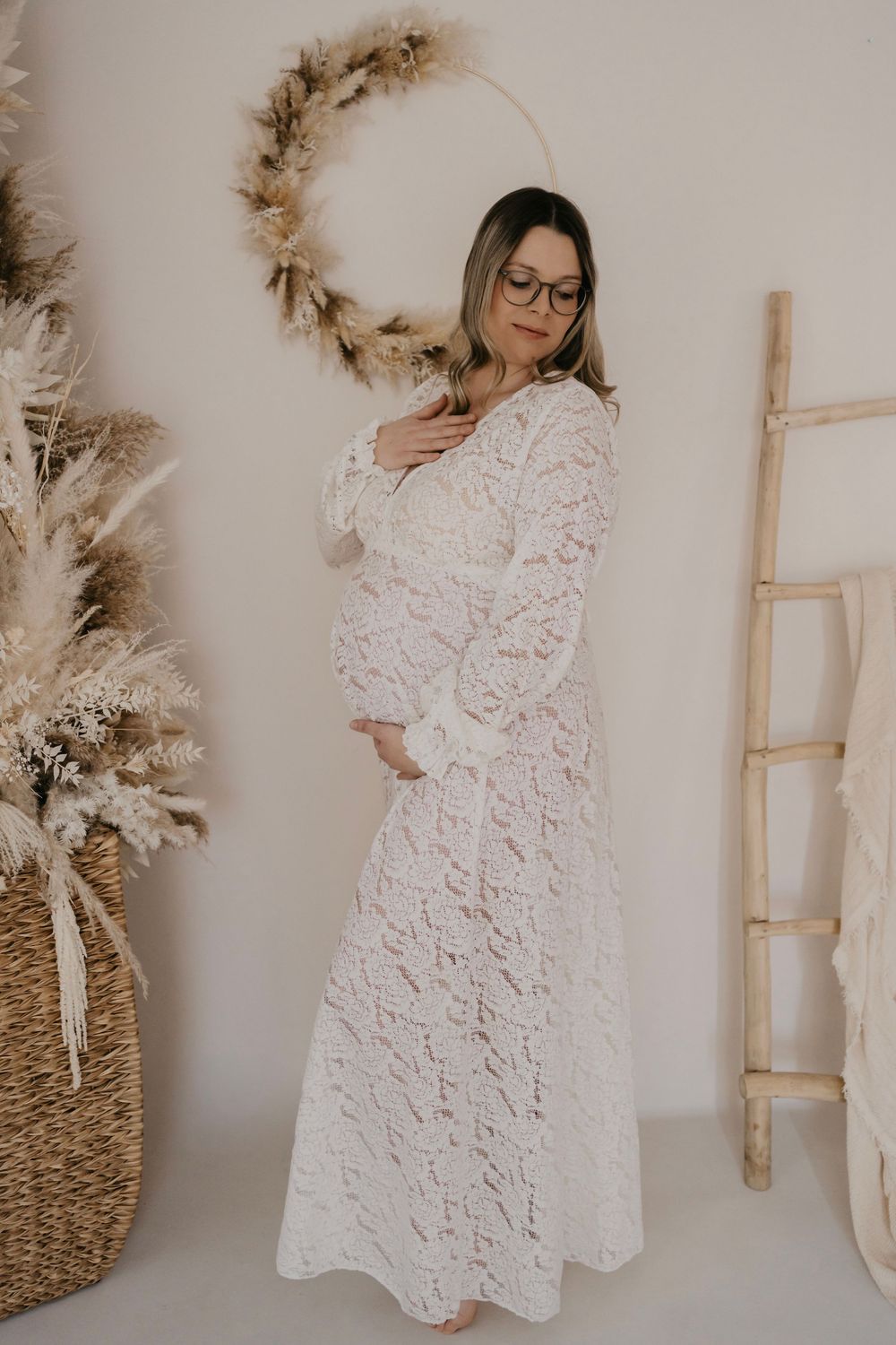 A white lace maternity dress styled with boho decor including pampas grass wreath and wooden ladder against neutral wall.