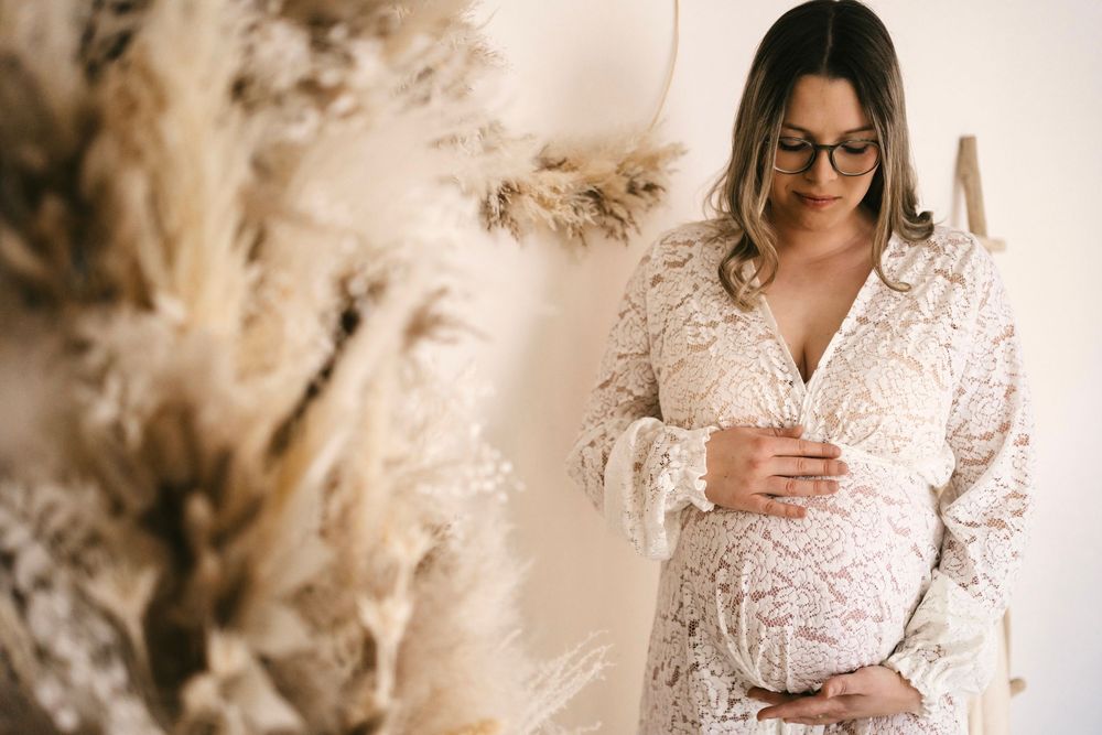 A pregnant woman in a white lace dress and glasses checks her phone while standing near decorative pampas grass.