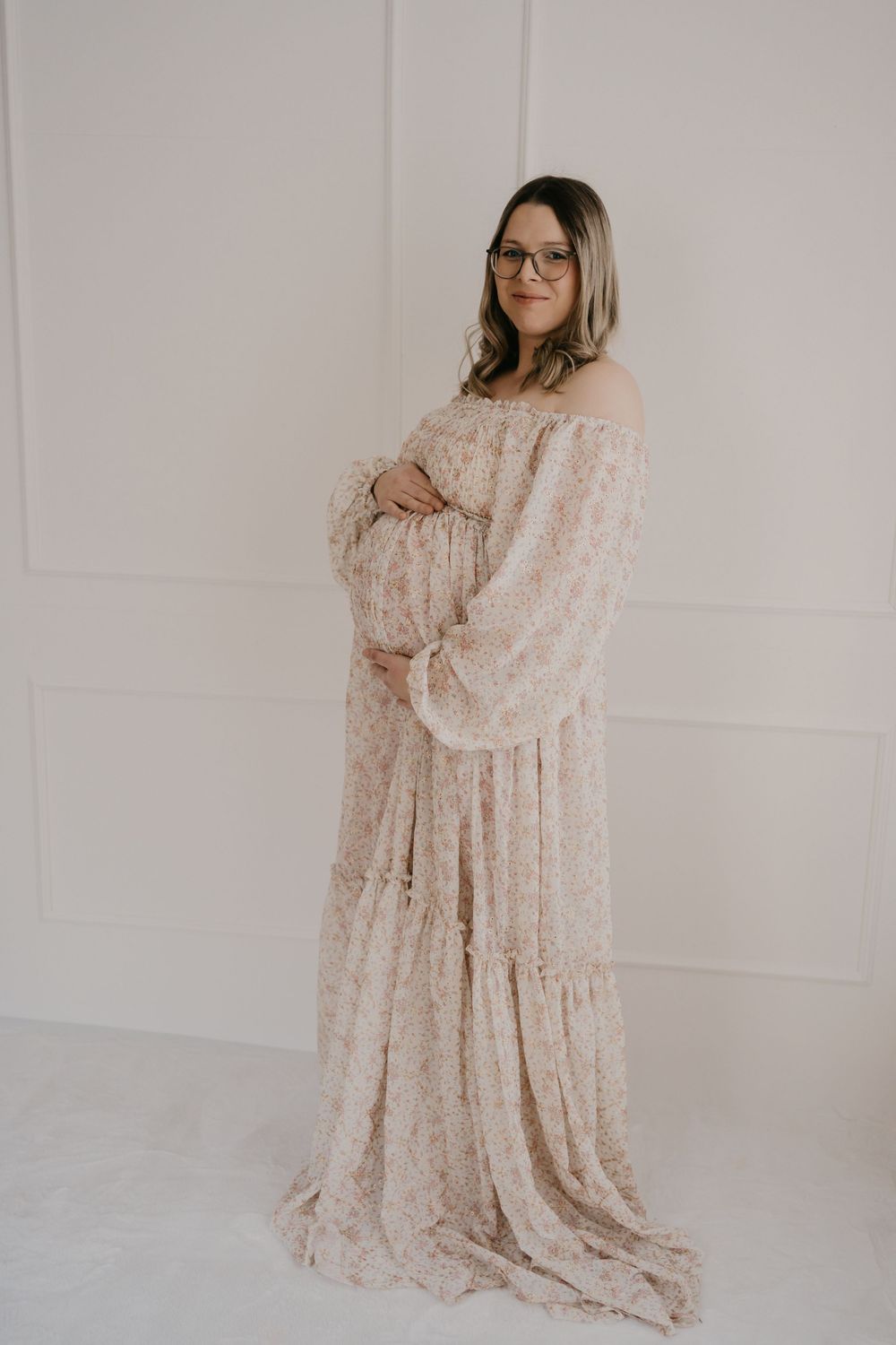 A person in glasses wearing a long flowy beige floral maternity dress stands against a white wall.