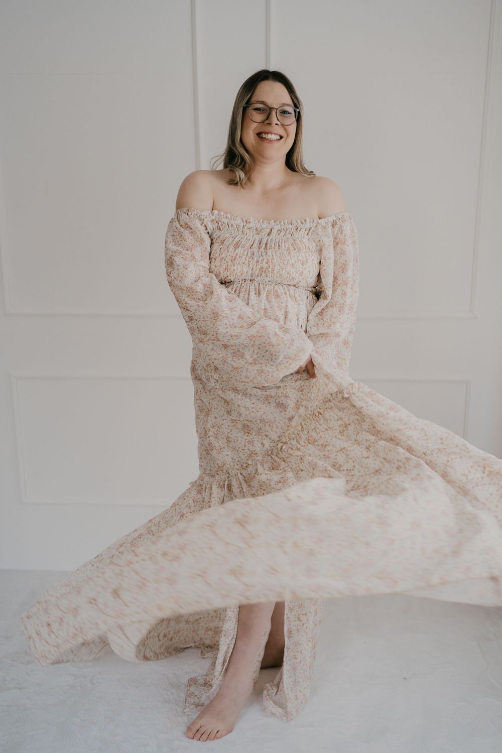 Floral off-shoulder dress with flowing fabric twirls in a romantic dance-like motion against a white wall backdrop.