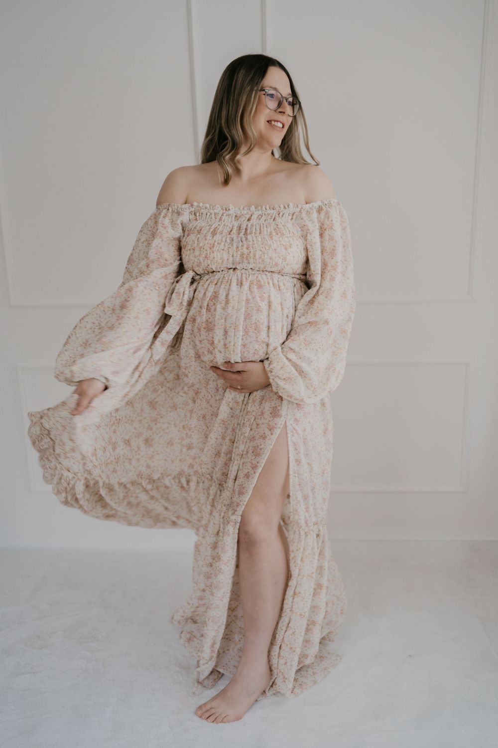 Expectant mother wearing flowy beige off-shoulder maternity dress with floral pattern poses barefoot against white wall.