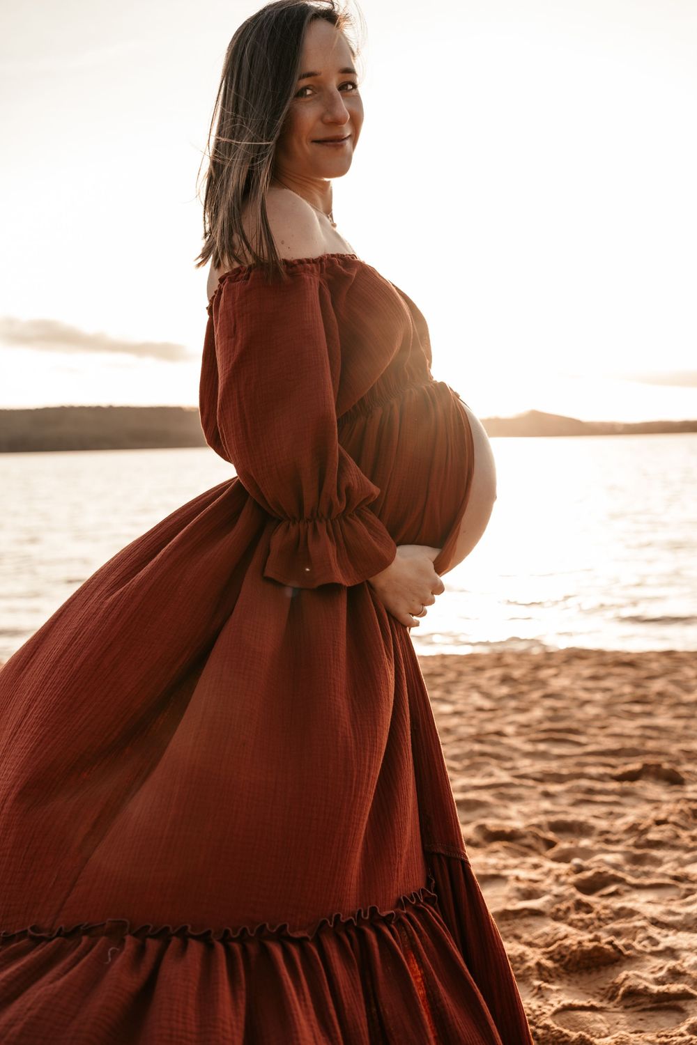 A pregnant woman in a flowing rust-colored dress poses on a sandy beach at sunset.