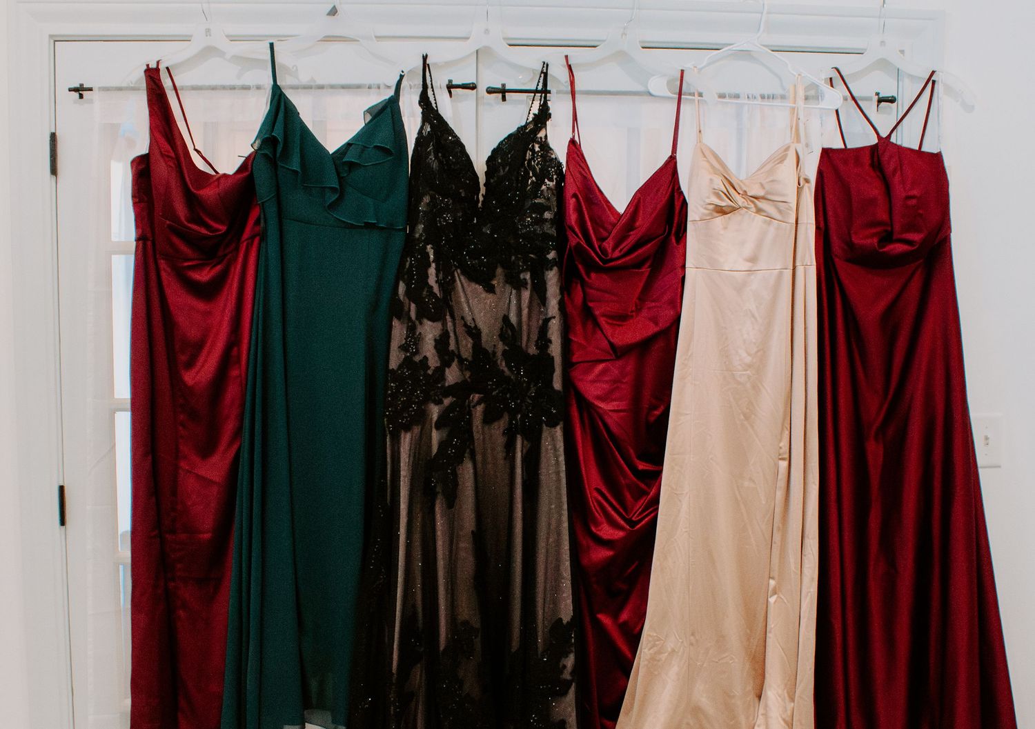 Five elegant formal gowns in red, green, black, nude, and burgundy hanging on a white door.