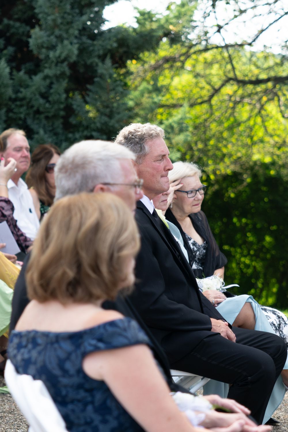 Wedding guests seated outdoors in formal attire at a garden ceremony with lush greenery in the background.