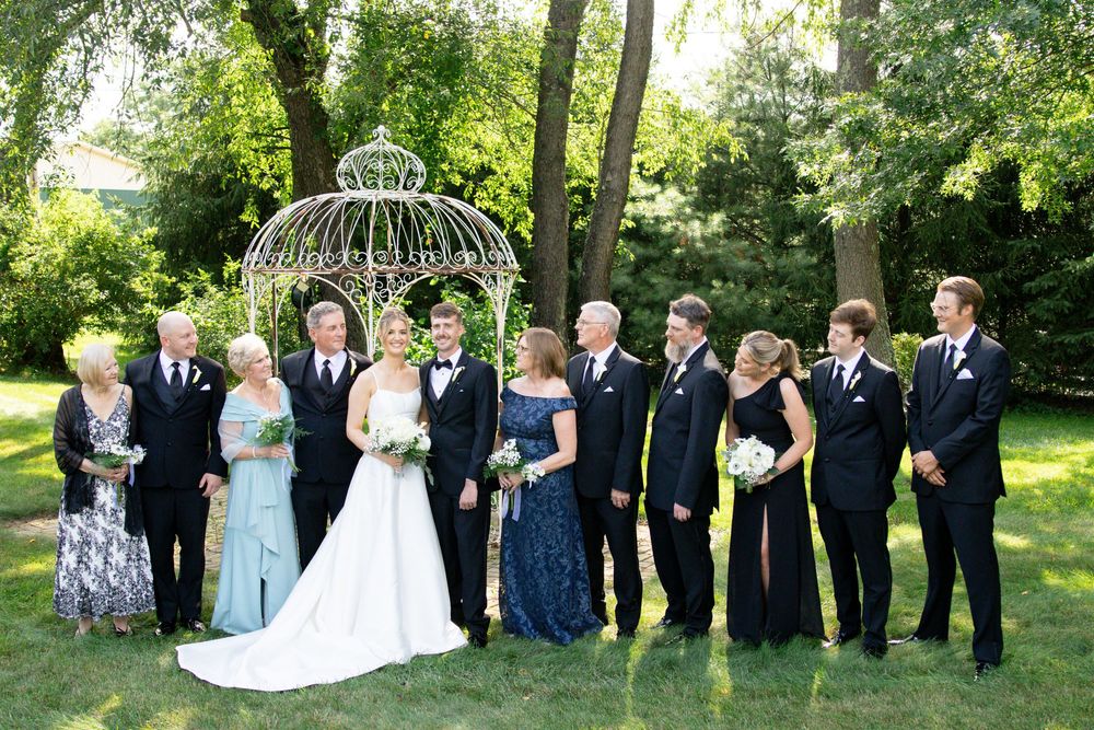 Wedding party lined up in formal black and white attire against a backdrop of green trees and gardens.