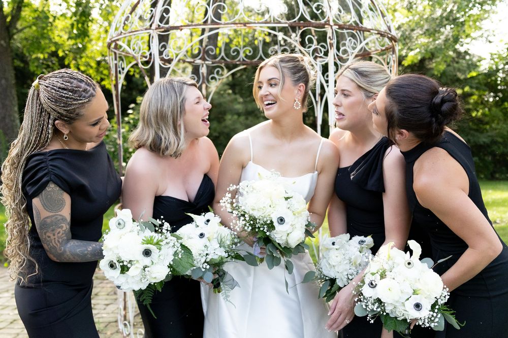 Wedding party members in black and white attire share joyful moments outdoors with white bouquets.
