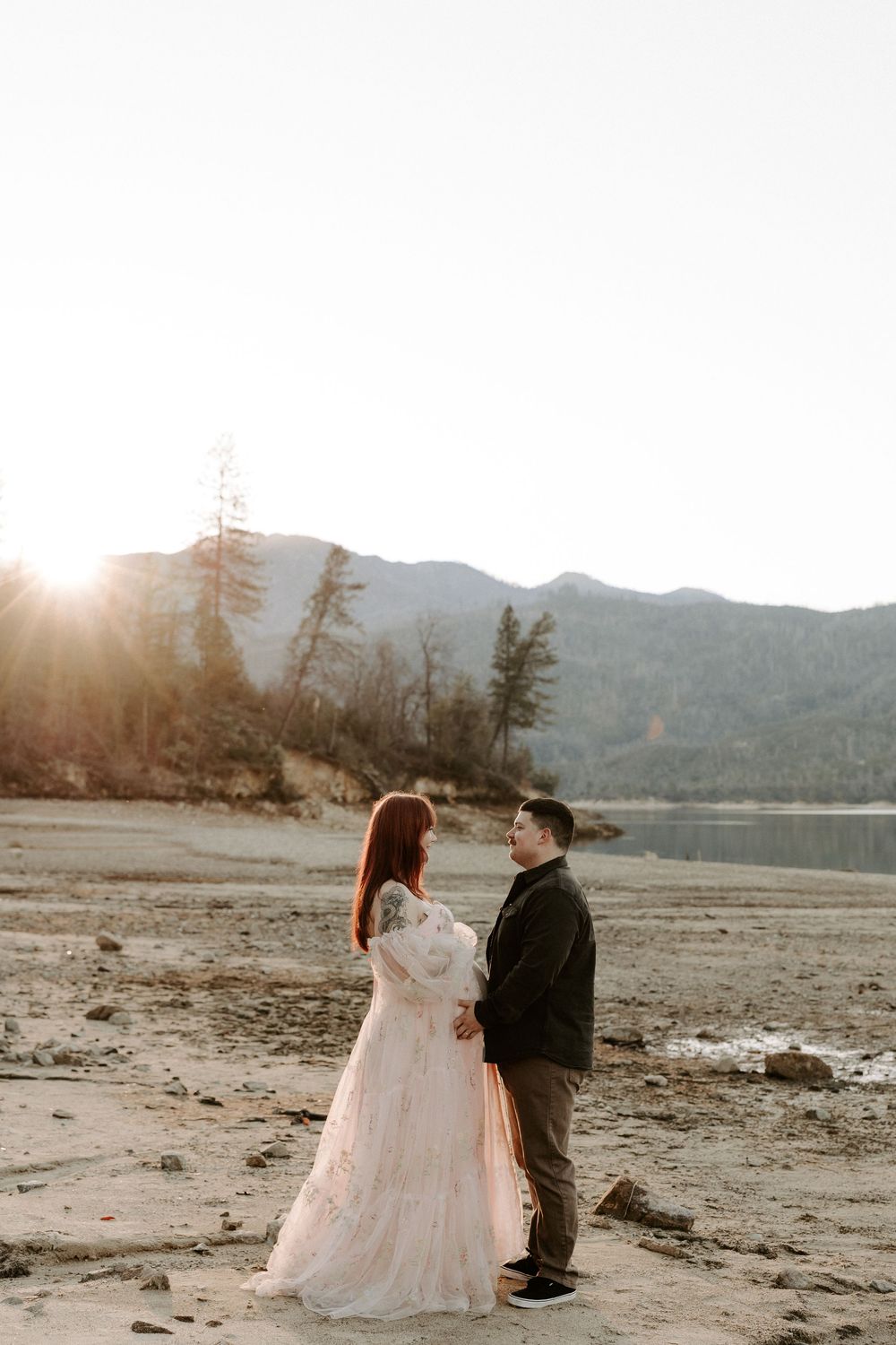 A couple shares an intimate moment on a sandy beach at sunset with mountains and trees in the background.
