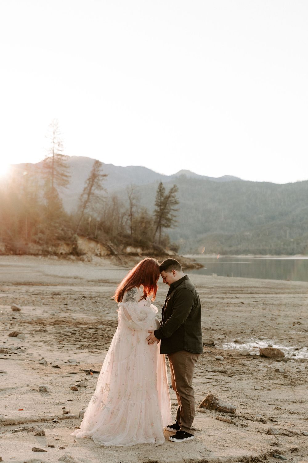 A couple in wedding attire shares intimate moments at sunset on a beach with mountains in the background.