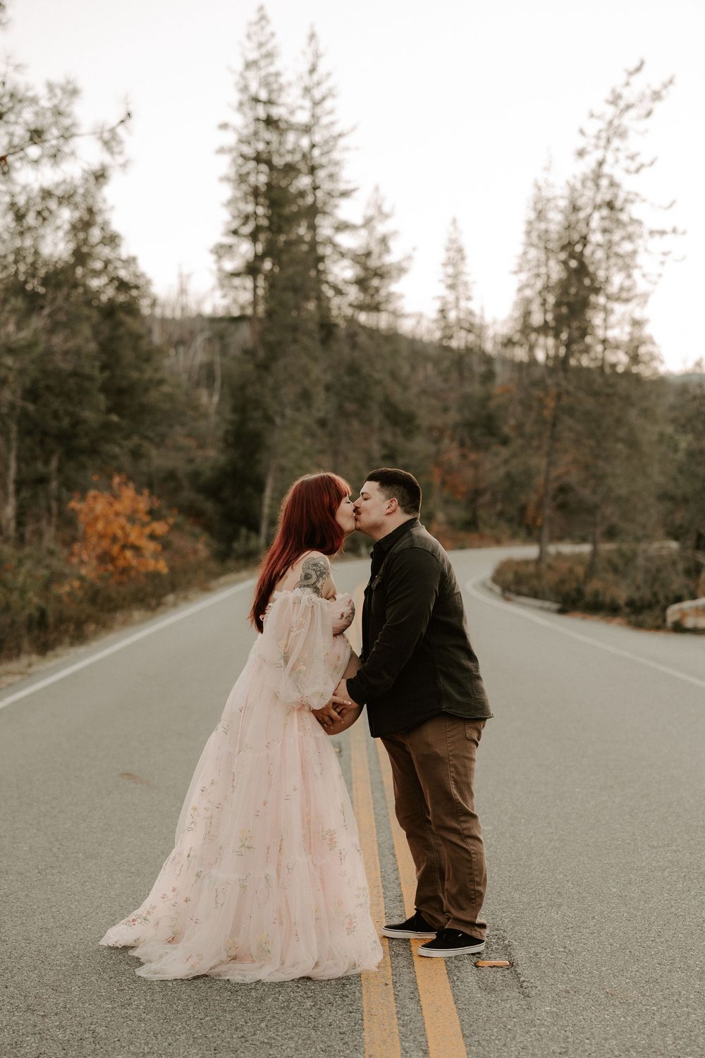 A romantic couple shares a tender moment on an empty road surrounded by tall evergreen trees and autumn foliage.