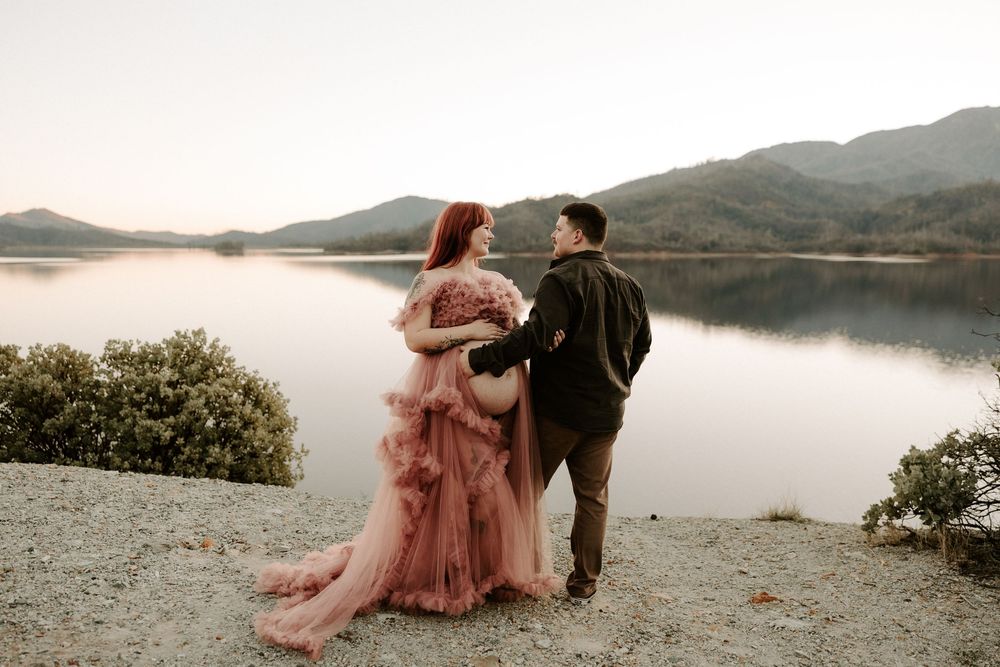 A romantic couple stands by a serene lake at sunset with mountains in the background, creating a dreamy atmosphere.