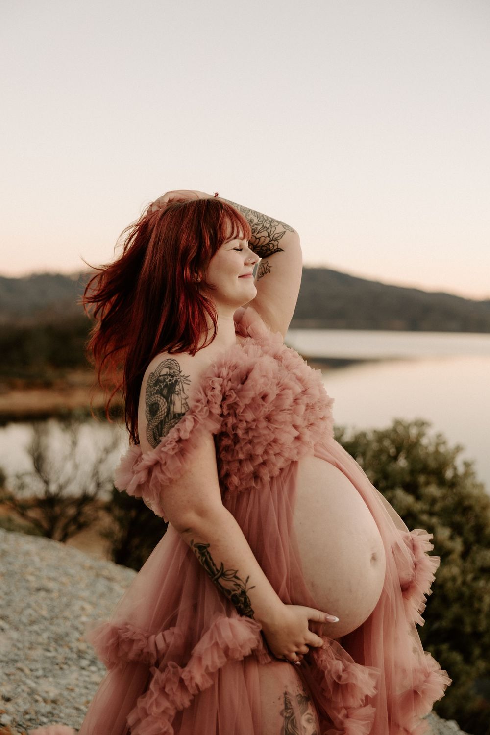 Maternity photo of woman in flowing pink tulle dress posing by lakeside at sunset.