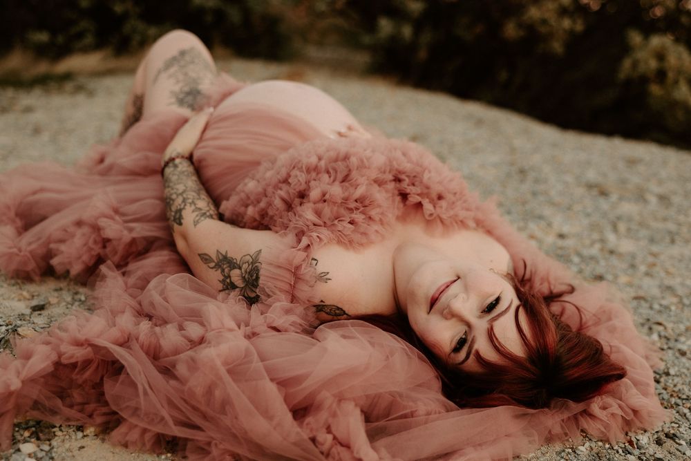 A person in a flowing dusty rose gown lies gracefully on the ground in an artistic outdoor photoshoot.