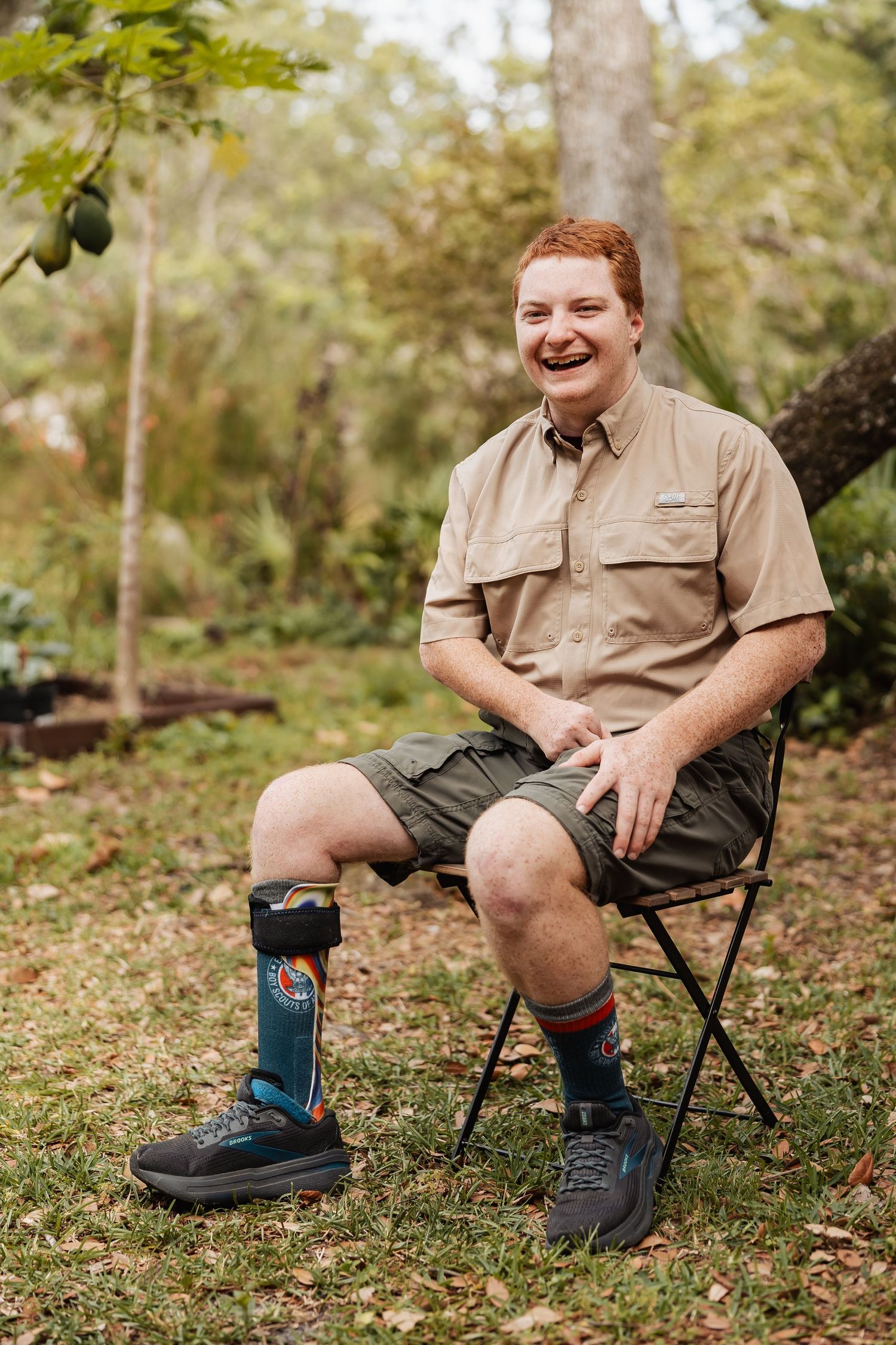 Person wearing khaki shorts and mismatched socks sits on a folding chair in an outdoor wooded setting.