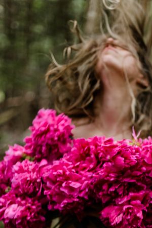 A person with windswept hair poses behind vibrant pink flowers in a garden setting.