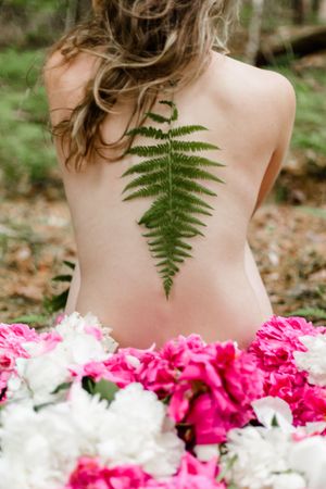 Artistic nude portrait with fern leaf and pink and white flowers creating ethereal natural composition.