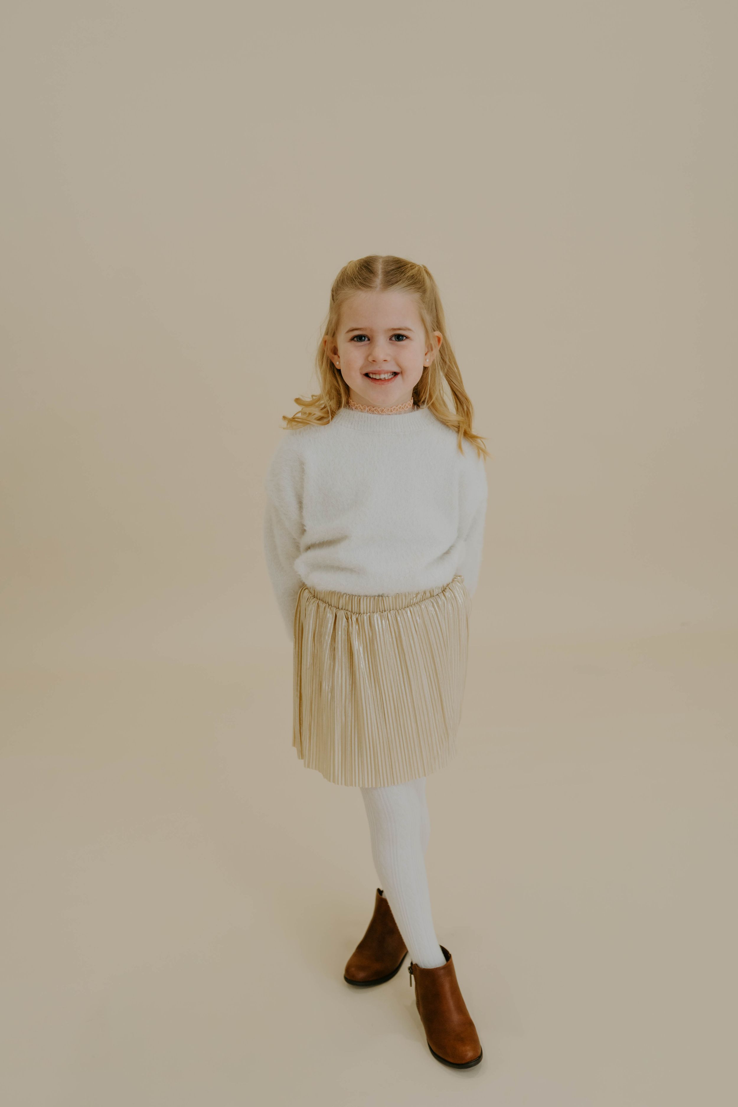 Young girl in white sweater and pleated skirt poses against beige backdrop.