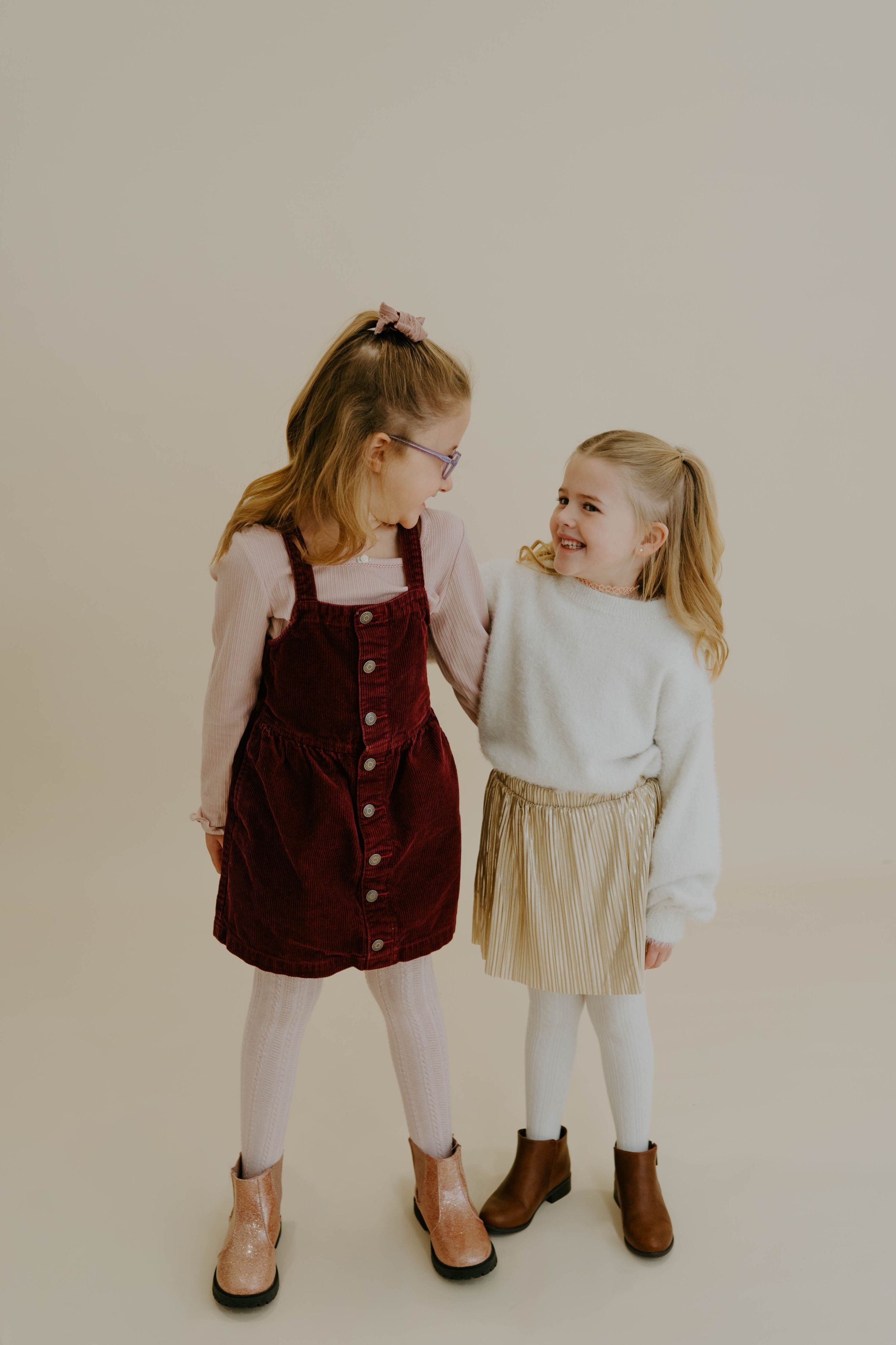Two children in coordinating outfits with a burgundy corduroy dress and cream sweater with gold skirt pose together.