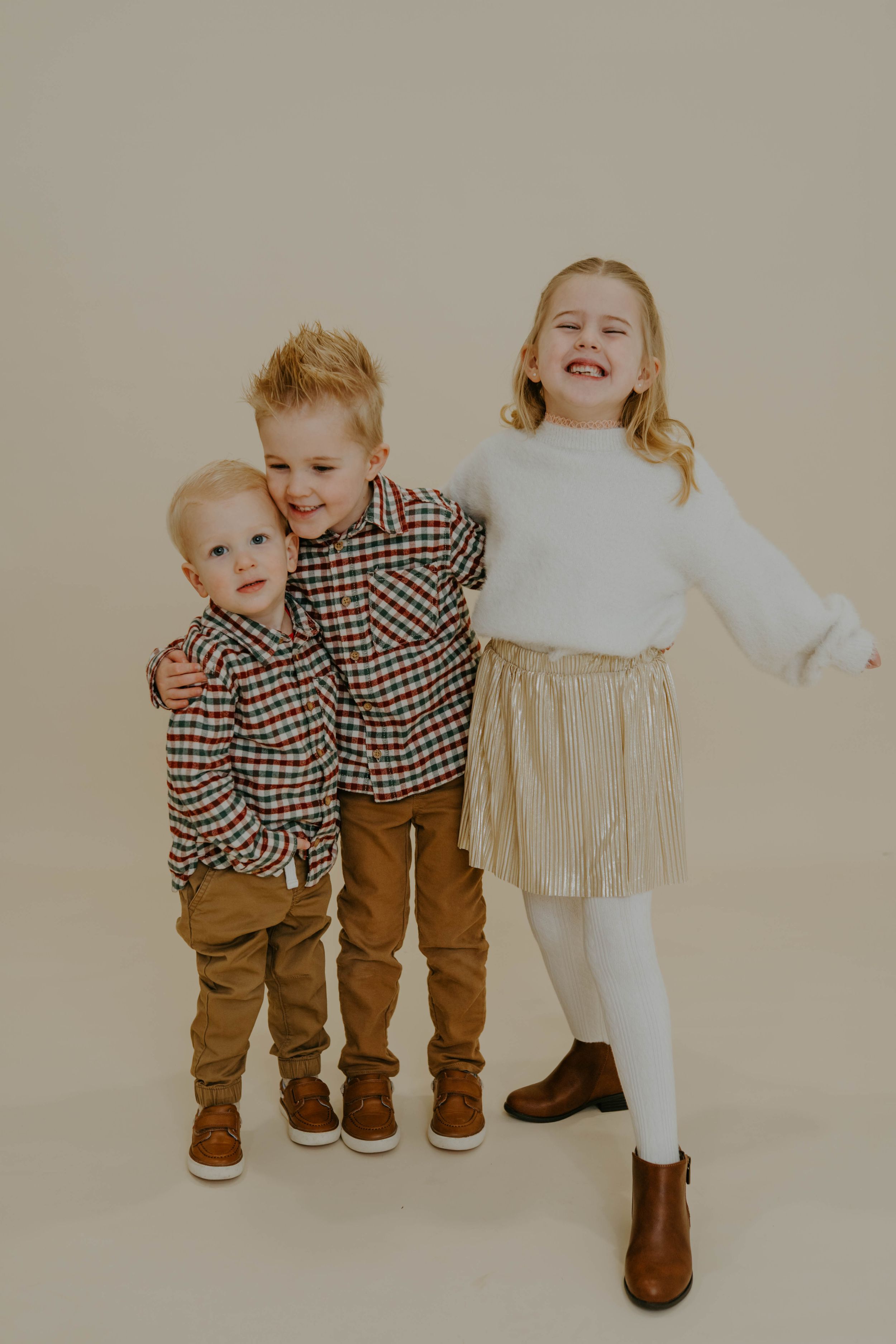 Young children posing together in matching plaid shirts and brown pants against neutral wall.