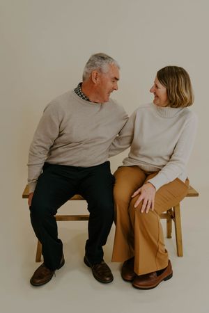 Two people in casual attire sitting together on a wooden bench against a neutral background.