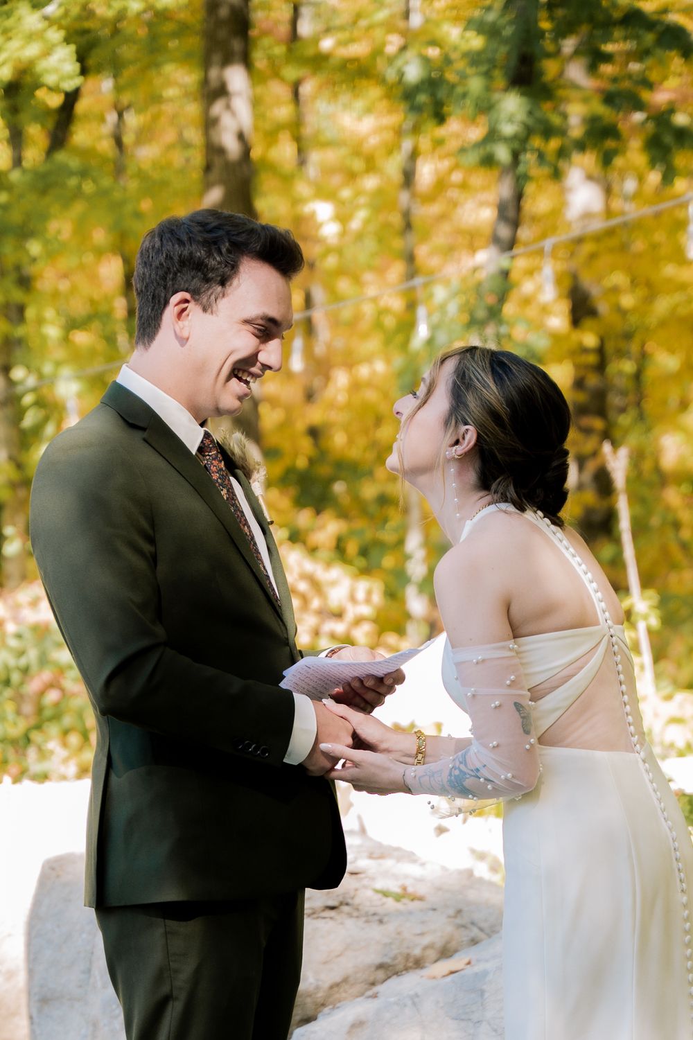 A couple shares a joyful moment laughing together during their winter wedding ceremony in a snow-covered forest setting.