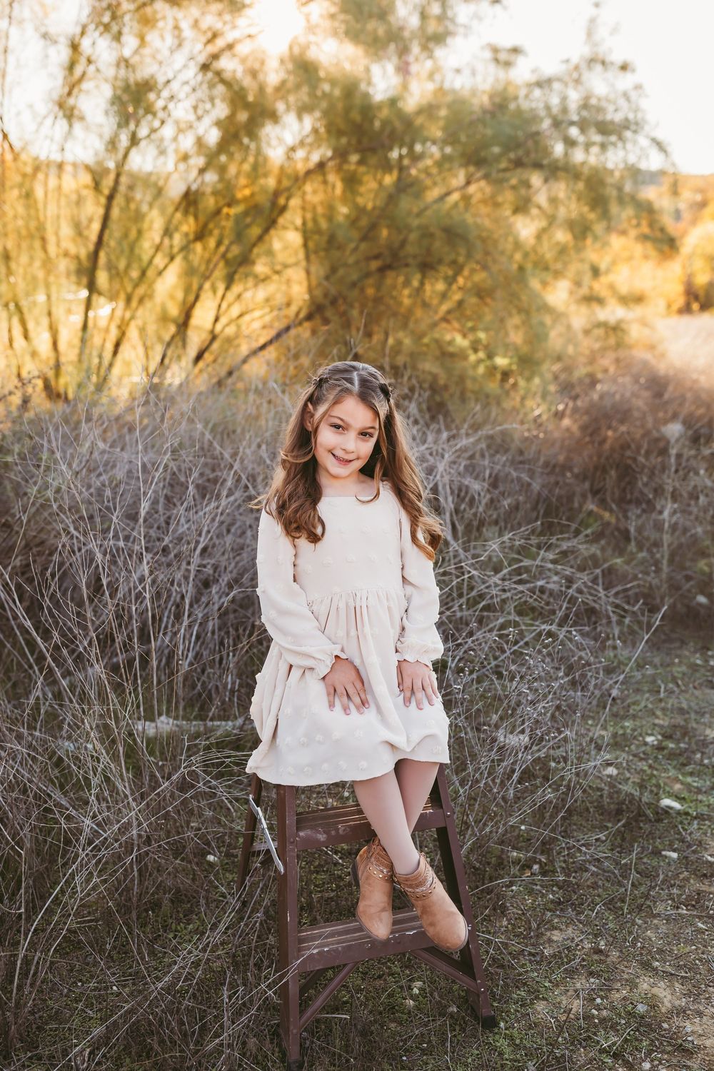 A person in a cream-colored dress sits on a rustic wooden chair surrounded by desert brush at golden hour in beaumont ca