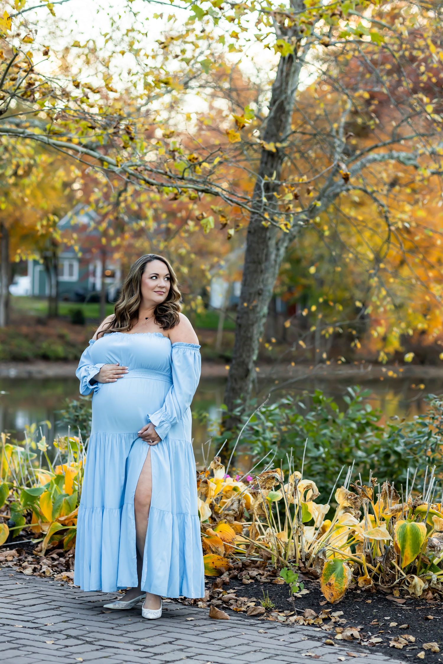 A person in a light blue dress poses outdoors among autumn trees and yellow flowers in a garden setting. RVA Maternity photograpger
