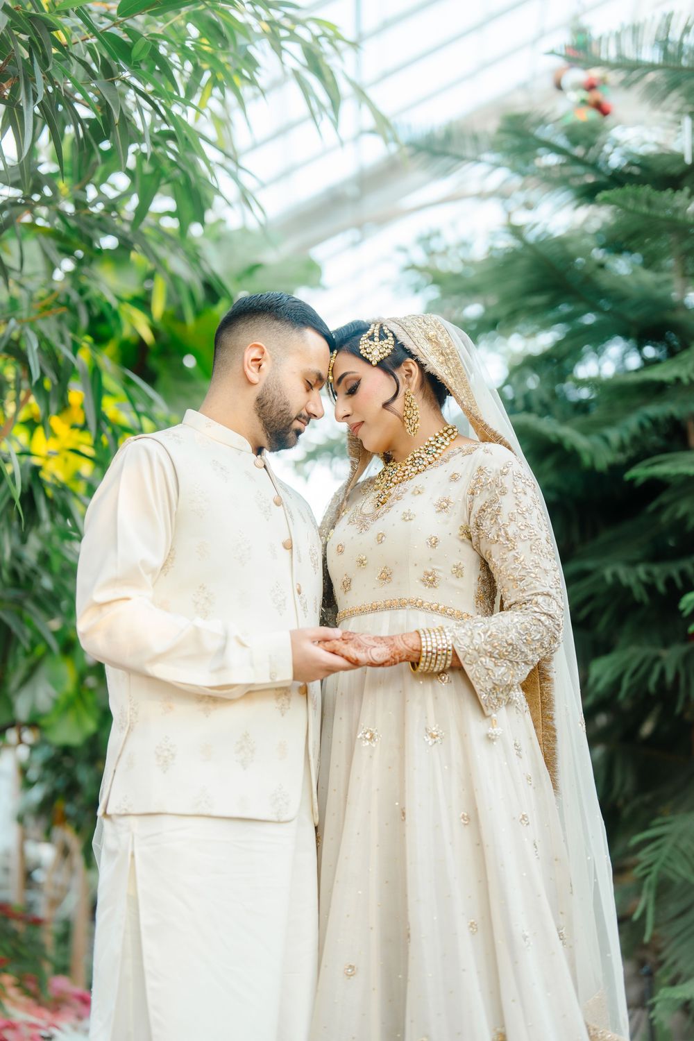 South Asian bride and groom in matching ivory wedding attire share an intimate moment in a greenhouse.