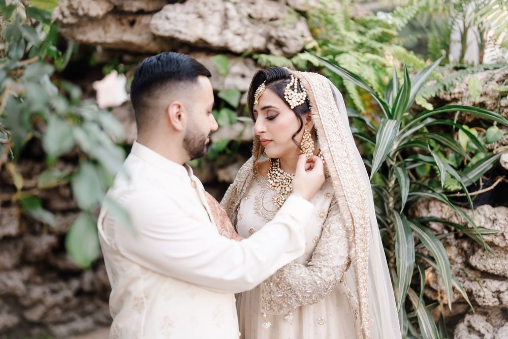 A groom in ivory sherwani tenderly adjusts his bride's jewelry in a garden setting during their wedding.