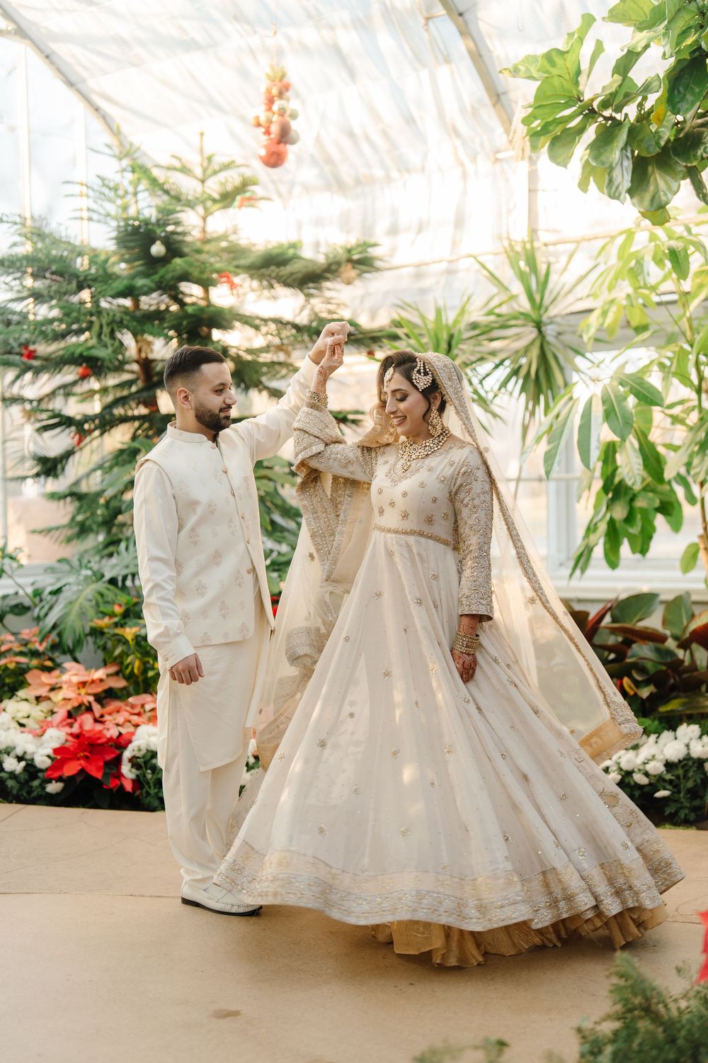 South Asian bride in ivory lehenga twirls joyfully as groom spins her in a lush botanical greenhouse.