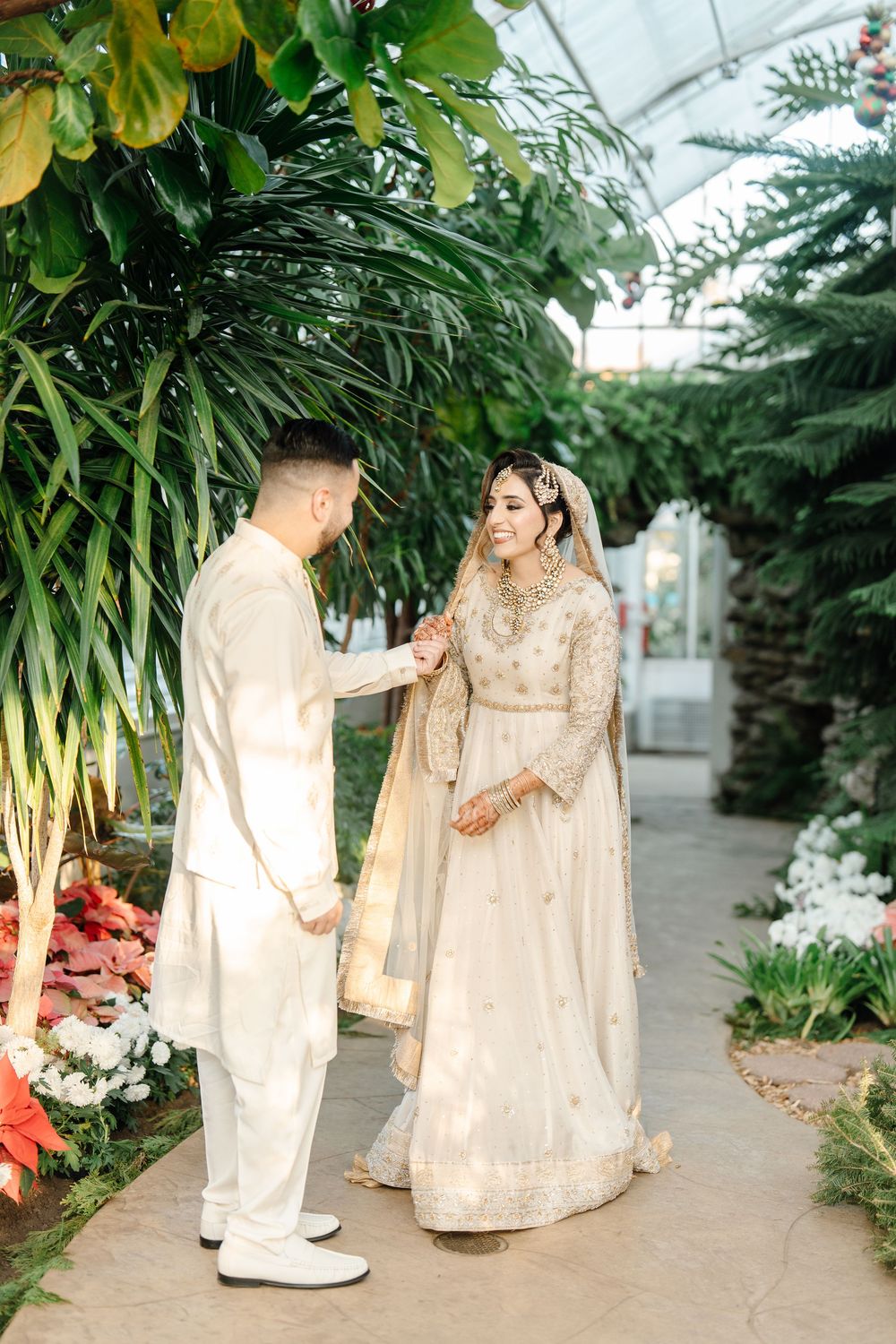 South Asian bride and groom holding hands in elegant ivory outfits inside botanical garden venue.