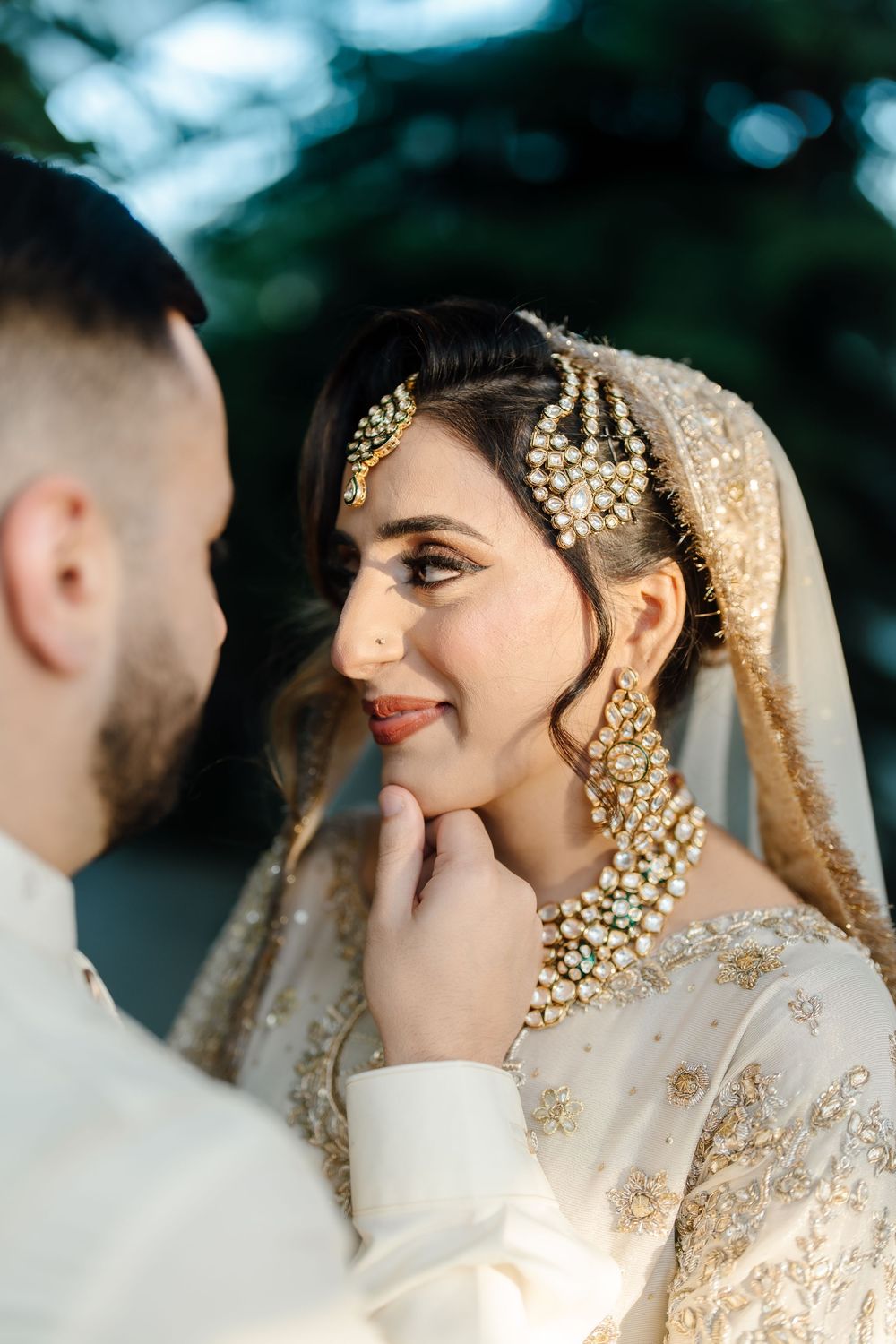 Groom tenderly touching South Asian bride's chin as she smiles in golden kundan bridal jewelry.
