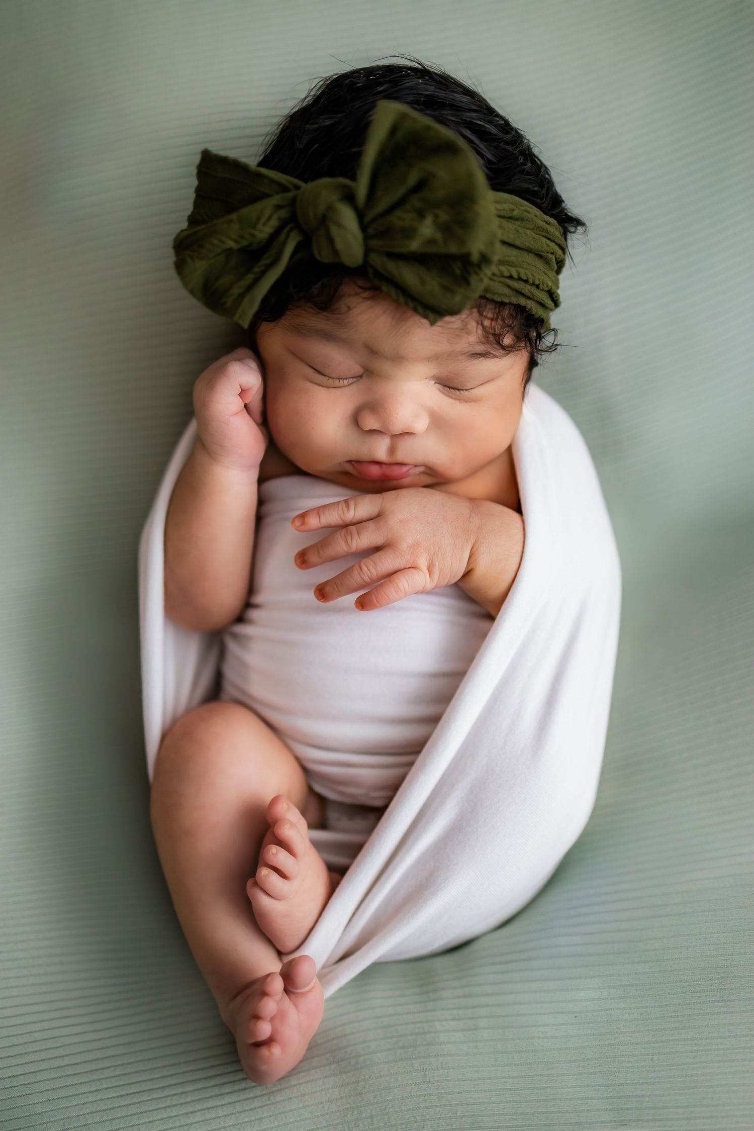 A sleeping newborn baby wrapped in white fabric with a green headband lies on a soft gray surface. Newborn photographer in Richmond, VA