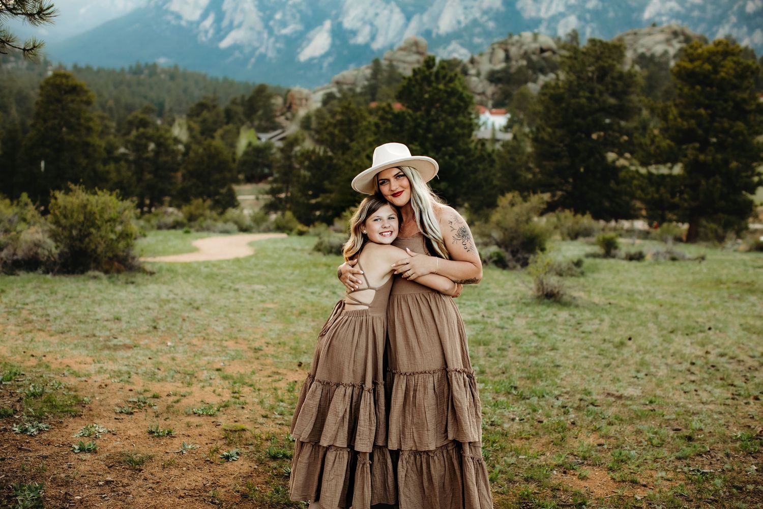 A couple in matching brown dresses poses in a scenic mountain landscape with evergreen trees and peaks in the background.