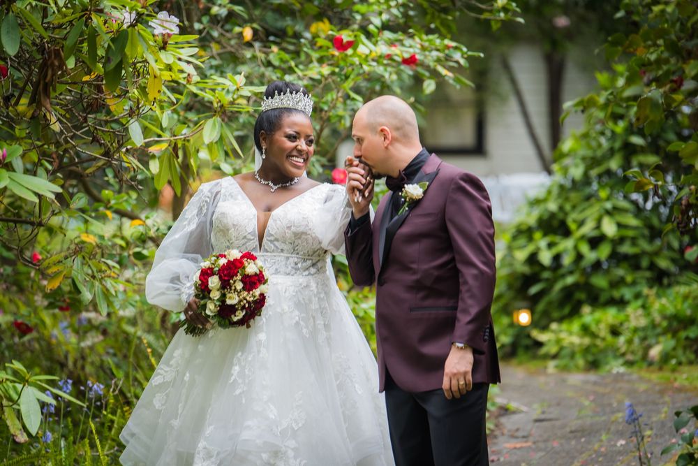 Couple in wedding attire stands together in a lush garden setting with white and red floral arrangements.