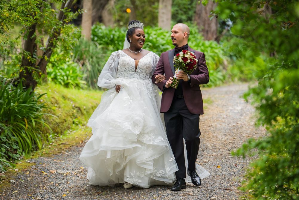 A couple walks down a lush garden path on their wedding day, with the bride in a flowing white gown and groom in burgundy suit.