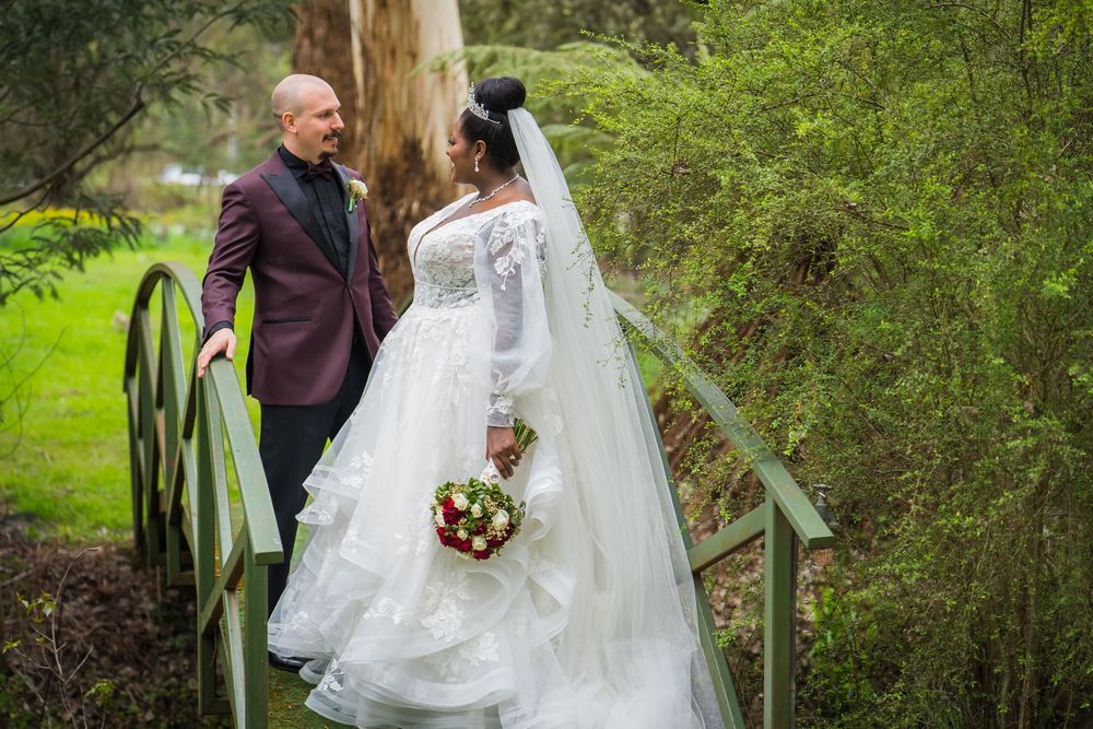 A couple in wedding attire stand together on wooden steps surrounded by lush greenery in a garden setting.
