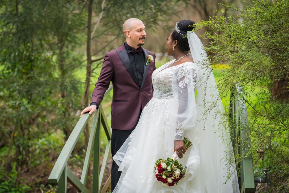 A couple in wedding attire stands on a garden bridge with greenery in the background, bride holding a bouquet.
