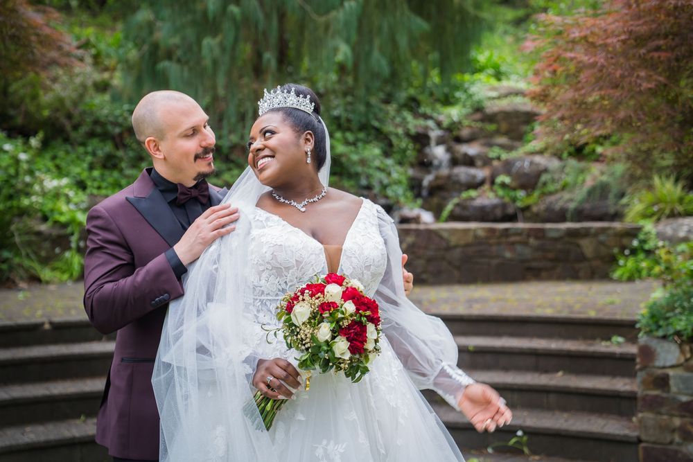 A couple shares a romantic moment in a garden setting while wearing wedding attire and holding a red bouquet.